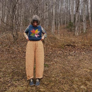 May include: A person wearing a denim vest with a colorful star design, a patterned long-sleeve shirt, and quilted tan pants. The person is standing in a forest setting, wearing blue shoes.