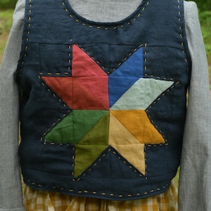 May include: A dark navy blue sleeveless vest featuring a large, multi-colored star appliqué on the front. The star is composed of red, blue, light blue, yellow, tan, and green fabric segments. The vest has visible yellow decorative stitching.