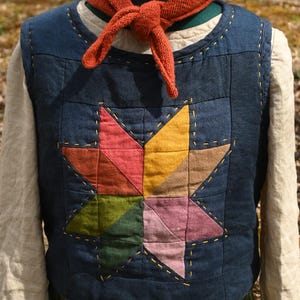 May include: A quilted vest in navy blue features a colorful star design. The star is made of various fabric pieces in shades of red, pink, yellow, green, and brown. A rust-colored scarf is tied around the neck of the mannequin.