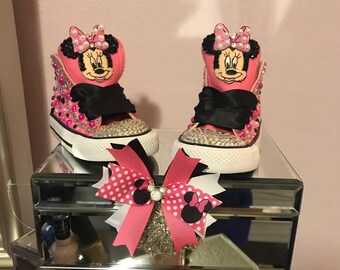 May include: Pink and black high-top sneakers with a Minnie Mouse design, rhinestones, and a black bow. The sneakers are on a mirrored surface with a pink and black bow.