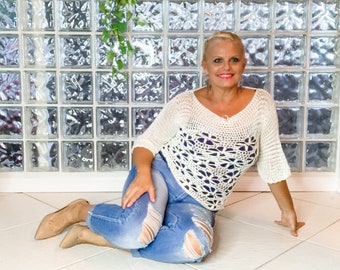 May include: A woman wearing a white crocheted top with a blue denim shirt underneath. She is sitting on a white tiled floor with her legs crossed.