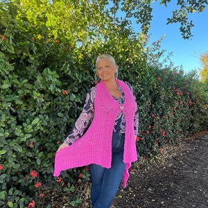 May include: A pink knitted vest with a shawl collar and a button closure. The vest is worn over a patterned blouse and blue jeans.