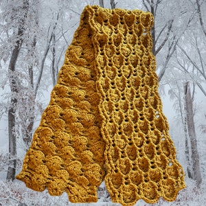Puede incluir: Una bufanda de crochet de color amarillo mostaza con un patrón texturizado y de encaje. La bufanda está doblada por la mitad y drapeada sobre un fondo nevado.