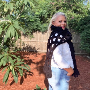 May include: A black crochet shawl with a white checkered pattern and fringe. The shawl is worn by a person with blonde hair and a white shirt.