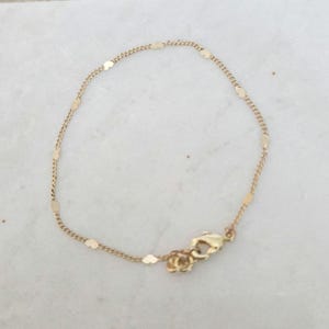 May include: Delicate gold bracelet featuring a chain with small, flat, rectangular charms. The bracelet has a lobster clasp closure. The jewelry is displayed on a white marble surface. A simple and elegant accessory.