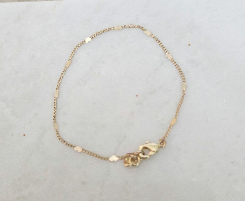 May include: Delicate gold bracelet featuring a chain with small, flat, rectangular charms. The bracelet has a lobster clasp closure. The jewelry is displayed on a white marble surface. A simple and elegant accessory.