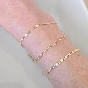 May include: Three gold bracelets of varying designs are displayed on a wrist. One bracelet features small, round charms, another has an oval link chain, and the third has a series of rectangular charms. The bracelets are made of a shiny, yellow metal.