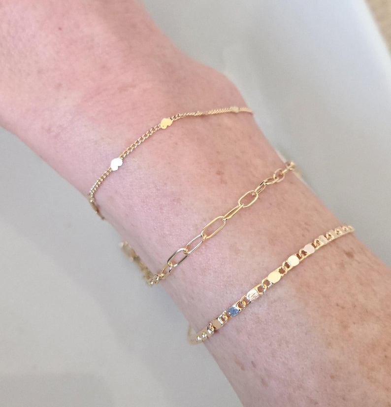 May include: Three gold bracelets of varying designs are displayed on a wrist. One bracelet features small, round charms, another has an oval link chain, and the third has a series of rectangular charms. The bracelets are made of a shiny, yellow metal.