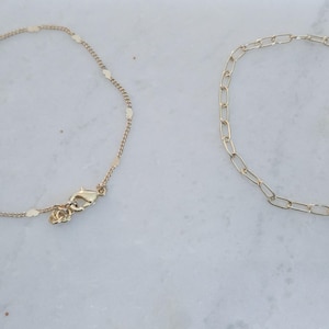 May include: Two gold-colored anklets. One features small, heart-shaped charms, while the other has a chain link design. Both have a lobster clasp closure and are displayed on a white marble surface.