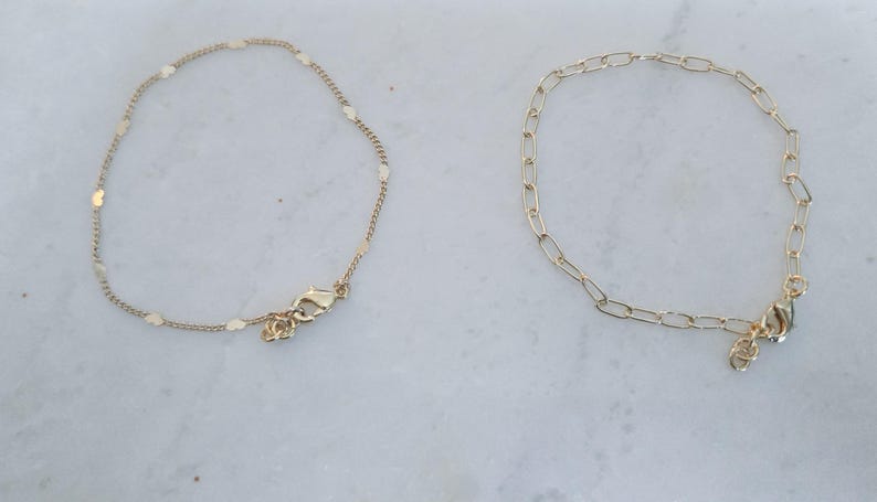 May include: Two gold-colored anklets. One features small, heart-shaped charms, while the other has a chain link design. Both have a lobster clasp closure and are displayed on a white marble surface.