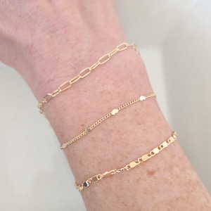 May include: Three gold bracelets are displayed on a wrist. The top bracelet has an oval link chain. The middle bracelet has small, round charms. The bottom bracelet has flat, rectangular links. The bracelets are delicate and shiny.