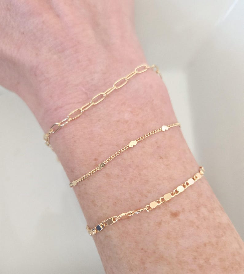 May include: Three gold bracelets are displayed on a wrist. The top bracelet has an oval link chain. The middle bracelet has small, round charms. The bottom bracelet has flat, rectangular links. The bracelets are delicate and shiny.