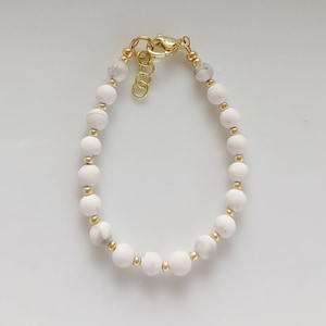 May include: A white beaded bracelet with gold accents. The bracelet is made of round, white beads with a subtle marbled pattern. The beads are strung on a gold chain with a gold clasp.