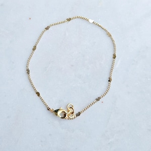 May include: Delicate gold-colored bracelet featuring a fine chain interspersed with small, flat, oval-shaped charms. The bracelet has a lobster clasp closure. The jewelry is likely made of gold-plated metal.