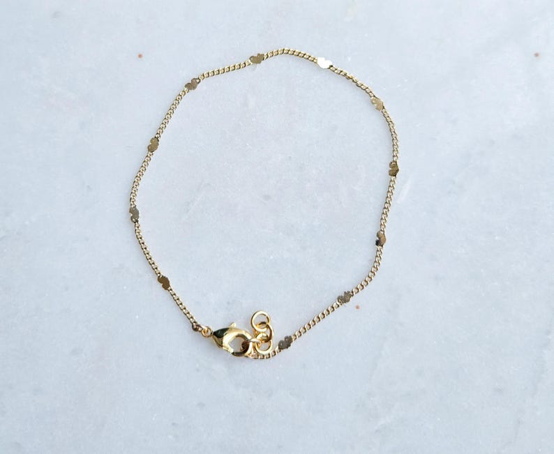 May include: Delicate gold-colored bracelet featuring a fine chain interspersed with small, flat, oval-shaped charms. The bracelet has a lobster clasp closure. The jewelry is likely made of gold-plated metal.