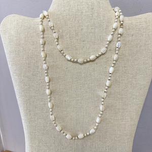 May include: A long, layered necklace featuring off-white, oval-shaped beads interspersed with smaller, round beads. The necklace is draped on a neutral-toned mannequin, showcasing its length and design. The beads have a subtle sheen.