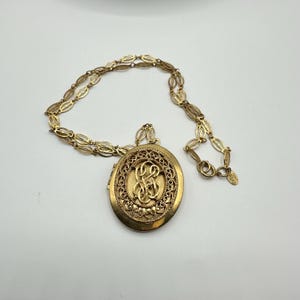 May include: A gold locket necklace with an oval-shaped locket featuring a delicate filigree design and an engraved monogram. The chain is made of linked oval shapes.
