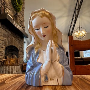 May include: A ceramic statue of a woman with blonde hair and a blue robe, hands clasped in prayer. The statue has gold accents and is set on a wooden surface. The background includes a fireplace and a decorative light fixture.