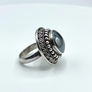 May include: A silver ring featuring a large, smooth, dark gray, oval-shaped stone set in the center. The setting is decorated with a beaded border and an outer band of engraved scrollwork patterns, creating a detailed, textured metal finish.