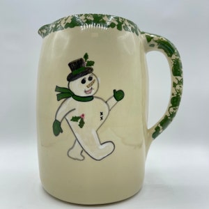 May include: A cream-colored ceramic pitcher with a green handle and green splatter design. The pitcher features a hand-painted snowman with a black top hat, green scarf, and green mittens.