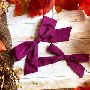 May include: Two plum-colored fabric bows, one large and one small, are displayed on a light wood-grain surface. The bows are tied in the center, with long, flowing tails. Autumn leaves and decorative branches surround the bows.
