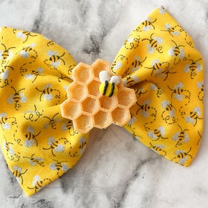 May include: A yellow fabric bow with a bee and honeycomb design. The bow is decorated with a small, black and yellow bee and a golden honeycomb.