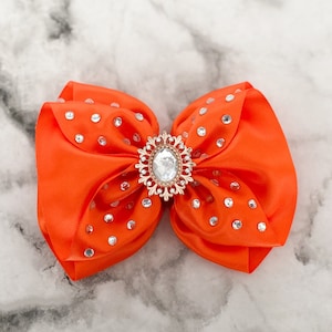 May include: Bright orange satin bow with a gold-toned, rhinestone-studded centerpiece. The bow is embellished with small, sparkling rhinestones. This hair accessory is perfect for adding a touch of glamour.
