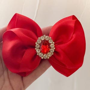 May include: A red satin bow with a large, clear rhinestone in the center. The rhinestone has a red center.