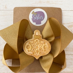 May include: A gold glitter hair bow with a beige, glittery, embossed ornament in the shape of a Christmas tree ornament. The ornament has a snowflake design.