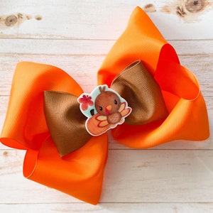 Könnte beinhalten: Orange-braunes Haarbändchen mit einem Truthahn-Applik. Der Truthahn ist braun mit einem roten Kehllappen und einem weißen Gesicht. Das Band ist aus Grosgrainband.