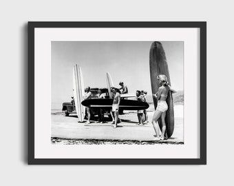 Puede incluir: Un grupo de personas se encuentran en una playa con tablas de surf. Una mujer en traje de baño sostiene una tabla de surf frente a ella. El grupo está de pie frente a un coche antiguo.
