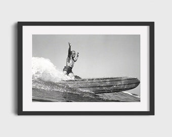 May include: A black and white photograph of a surfer riding a wave on a longboard. The surfer is wearing a swimsuit and has their arms raised in the air.
