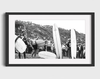 May include: Black and white photograph of five surfers standing on a beach with their surfboards. The surfers are wearing swimsuits and have numbers on their chests. There are many people in the background watching the surfers.