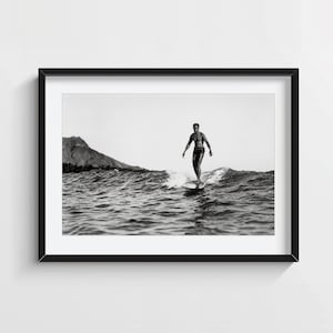 Op de afbeelding: Zwart-wit ingelijste print van een surfer die op een golf rijdt. De surfer staat op een surfboard in de oceaan, met een berg op de achtergrond. De afbeelding zit in een zwart frame.