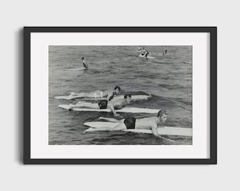May include: A black and white photograph of five people lying on surfboards in the ocean. The people are all wearing swimsuits and are looking towards the camera. The water is choppy and there are waves in the background.