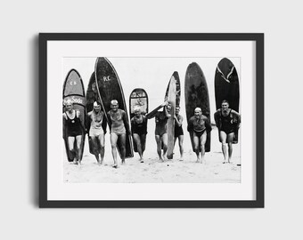 May include: A black and white photograph of eight men carrying surfboards on a beach. The surfboards have various markings, including "C.B. Soda" and "R.C."