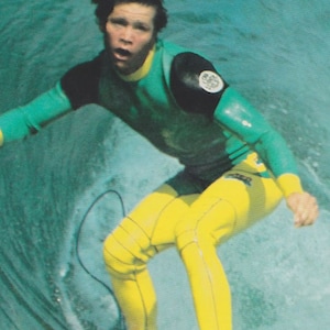 May include: A surfer wearing a green and yellow wetsuit with a black patch on the shoulder. The surfer is riding a wave and has a determined expression on their face.
