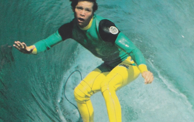 May include: A surfer wearing a green and yellow wetsuit with a black patch on the shoulder. The surfer is riding a wave and has a determined expression on their face.