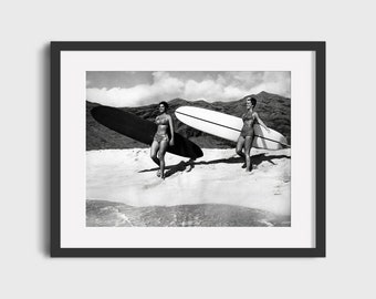 May include: Two women in swimsuits walk along a beach carrying surfboards. One surfboard is white and the other is black.