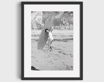 May include: A black and white photograph of a woman in a swimsuit carrying a surfboard on a sandy beach.