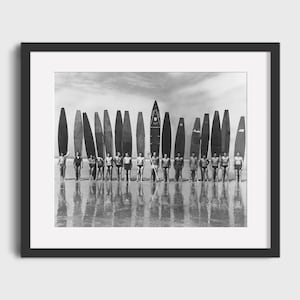 May include: A black and white photograph of a group of men standing on a beach with surfboards. The men are all wearing swimsuits and are holding their surfboards. The surfboards are all different sizes and shapes. The beach is sandy and the water is calm.
