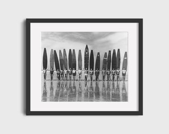 May include: A black and white photograph of a group of men standing on a beach with surfboards. The men are all wearing swimsuits and are holding their surfboards. The surfboards are all different sizes and shapes. The beach is sandy and the water is calm.