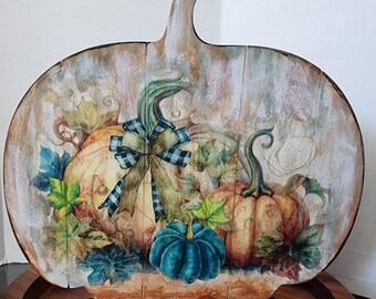 Peut inclure: Un support décoratif en bois en forme de citrouille avec un motif peint de trois citrouilles, dont une avec un nœud à carreaux bleu et blanc, et des feuilles d'automne. Le support est sur une base en bois.