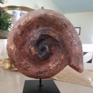 May include: A brown and gold spiral shell sculpture on a black metal stand. The shell has a textured surface and a swirling pattern.