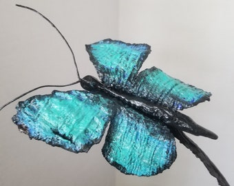 May include: A teal blue butterfly sculpture with black accents and a wire stem. The butterfly has a textured surface and is made of metal.