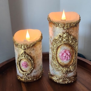 May include: Two decorative pillar candles with a floral design and gold accents. Each candle features a pink rose in a gold frame. The candles have a flickering LED flame and are placed on a wooden tray.