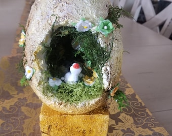 May include: A white ceramic egg with a moss-covered opening, revealing a small white bird figurine inside. The egg is decorated with white flowers and green leaves and sits on a gold base.