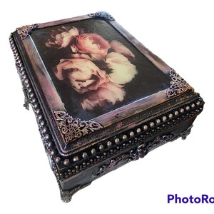 Puede incluir: Una caja de madera decorativa con un diseño floral en la tapa. La caja está pintada en tonos de negro, marrón y rosa. La tapa está adornada con un borde de cuentas plateadas y esquinas de metal ornamentadas.