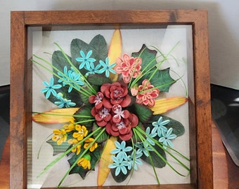 Puede incluir: Un arreglo floral enmarcado con una variedad de flores en tonos de azul, amarillo, rojo y naranja. Las flores están hechas de papel y están dispuestas en un patrón circular. El marco está hecho de madera y tiene un frente de vidrio.