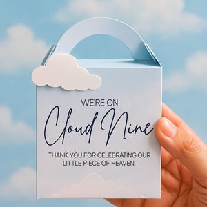 Puede incluir: Caja de regalo azul claro con adorno de nube blanca y asa. La caja presenta el texto "WE'RE ON Cloud Nine" y "THANK YOU FOR CELEBRATING OUR LITTLE PIECE OF HEAVEN" en una elegante escritura.